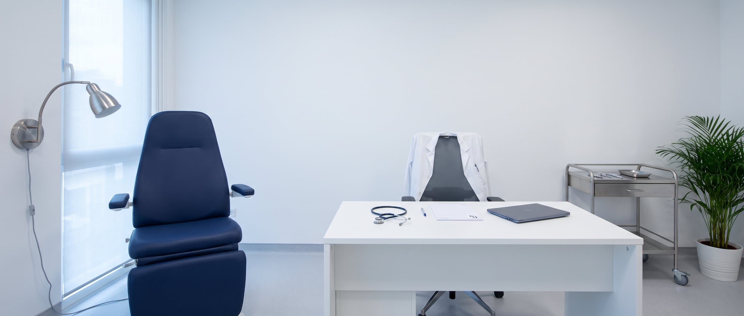 A bright consultation room: a navy treatment chair, a white desk with a stethoscope and prescription pad, and a white coat over the doctor’s chair